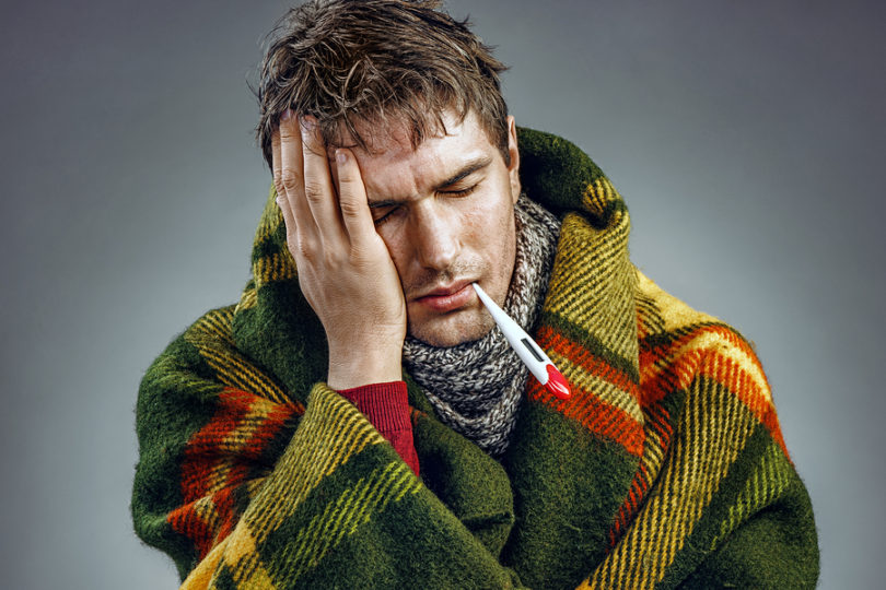 Photo of sick man wrapped in blanket with a thermometer in his mouth. 